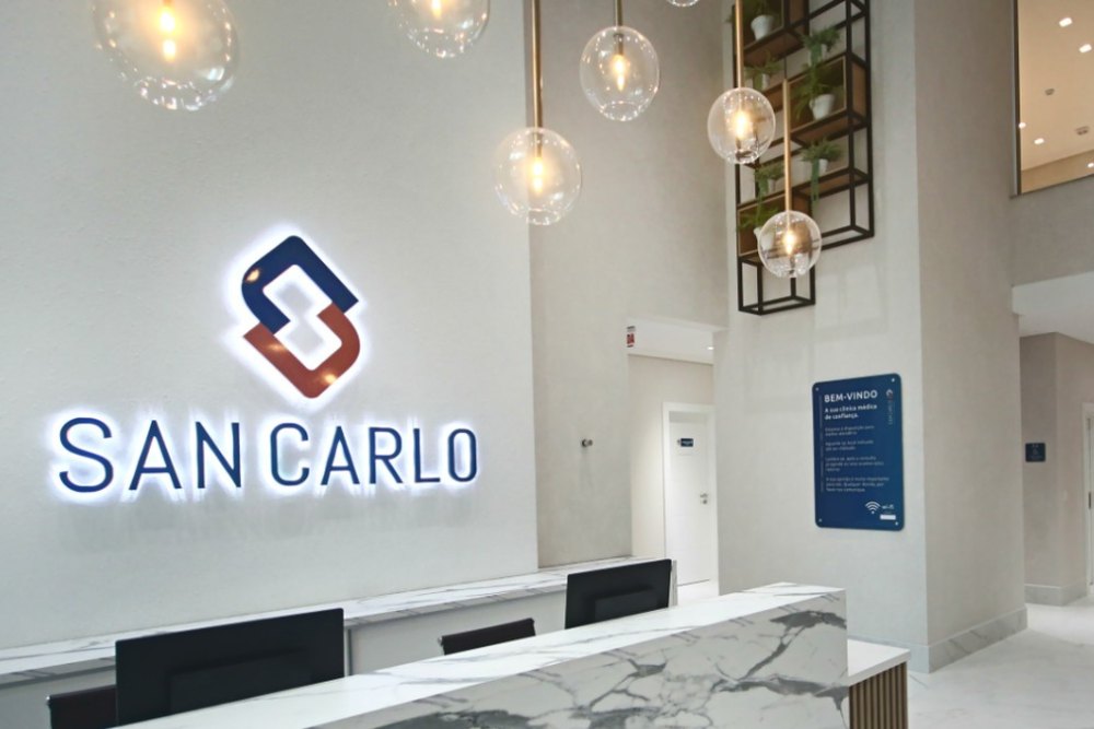 Recepção da Clínica San Carlo, em Balneário Camboriú, com o logotipo iluminado na parede e o balcão de atendimento
